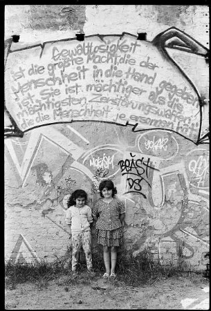 Zwei Mädchen vor einer Graffiti-Wand mit einem Zitat Mahatma Ghandis "Gewaltlosigkeit ist die größte Macht, die der Menschheit in die Hand gegeben ist. Sie ist mächtigerr als die mächtigsten Zerstörungswaffen, die die Menschheit je ersonnen hat."