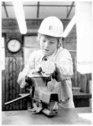 Junge mit Schutzhelm feilt an einem Werkstück , das in einen Schraubstock eingespannt ist (Altersgruppe 14-17)