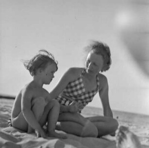 Frau in Bademode und nacktes Kind sitzen am Strand
