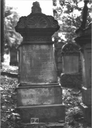 Grabstein 94 (Index: 99898869) Material: Sandstein Zustand: stark verwittert Maße HxBxT: 93x61x26 cm Beschreibung: aufgesockelte Stele, Giebelaufsatz mit Eckakroterien, Blüten- und Blattornamenten, unterer Sockel felsartig gearbeitet, Grabeinfassung Symbolik: Blüten- und Blattornamente: Grabstein 94
