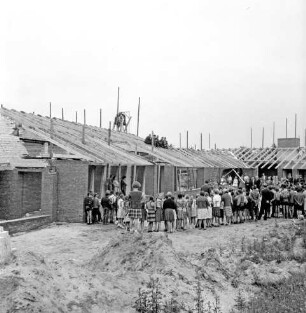 Volksschule: Neubau: Rohbau: Richtfest: vorn Sand- und Erdhaufen und Gestrüpp: dahinter Schülerhalbkreis: rechts dahinter Funktionsträger: hinten Pavillonbauten mit Richtkrone und Arbeitern auf Dachsparren