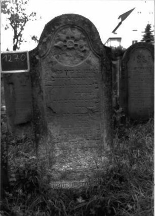 Grabstein 1270 (Index: 100104503) Material: Sandstein Zustand: stark verwittert Maße HxBxT: 180x68x20 cm Beschreibung: Stele, Konvexbogenabschluss, Schriftfeld eingetieft, im Bogenfeld plastische Blüte in rundem Medaillon, umlaufender winziger Zahnfries ? Symbolik: Blüte in Medaillon, Zahnfries ? Besonderheit: kulturhistorisch wertvoll / hist.-wiss. Gründe (Vorbeter, Lehrer): Grabstein 1270