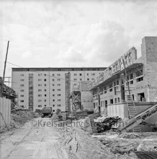Klosterbergen: Eichenbusch: rechts und links dreigeschossige Wohnblocksim Bau: in der Mitte Grünanlage: dazwischen Sandstraße mit Sandhaufen, Brettern und Baumaterial: rechts Leiter, Förderband, Ziegelstapel und Holzstangen zum Gerüstbau: hinten Mitte Lkw: dahinter achtgeschossiges Punkthaus nach Fertigstellung