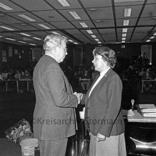 Manfred Samusch: Wiederwahl: in Stadtverordnetenversammlung: Gratulation durch Bürgervorsteherin Maria Heitmann: Tisch mit Akten und Mikrofon, Blumenstrauß: hinten Personen, 9. April 1984