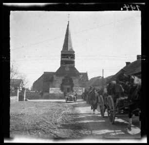 Unbekannte Ortschaft [Nordfrankreich?]: Pferdegespanne mit Wagenkarren auf einer unbefestigten Ortsstraße, im Hintergrund ein Personenkraftwagen mit offenem Verdeck und die Ortskirche