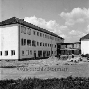 Ahrensburg: Hermann-Löns-Straße: Erweiterungsbau: vor Fertigstellung: Außenansicht: Herrichten des Vorplatzes: Arbeiter beim Verlegen von Betonplatten, Kleintransporter: hinten Bildmitte Verbindungstrakt, rechts Altbau