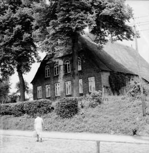 Groß Barnitz: Bauernhaus mit Reetdach: davor zwei Bäume und Vorgarten mit Hecke: vorn auf Gehweg Frau