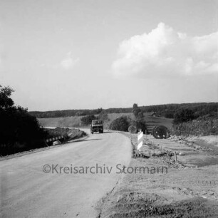 Straßenbau: Oldesloer Straße: Ausbau: rechts und links unbefestigte Randstreifen mit Absperrungen: rechts Kabelrollen: hinten Baufahrzeug: im Hintergrund Knicklandschaft
