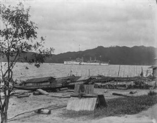 Am Hafen : Hausboote liegen am Strand trocken, im Hintergrund Schiff des Ostasiengeschwaders