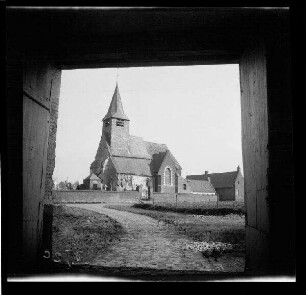 Tourmignies (Dep. Nord, Frankreich): "Kirche [Saint-Pierre-d'Antioche] von Tourmignies" mit Friedhof, im Vordergrund ein Feld (Seitansicht, Distanzaufnahme durch ein Hoftor)