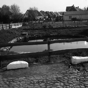 Dorfteich mit Grünanlage und Holzgeländer: davor weiß angemalte Findlinge: hinten Neubausiedlung mit Einfamilienhäusern und Gärten