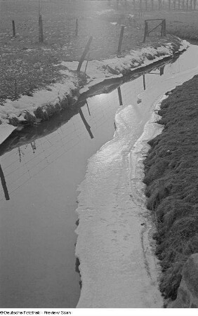 Ein vereister Bachlauf, am Ufer ein Zaun