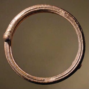 Massiver runder Armring von rundem Querschnitt mit übergreifenden Enden; an der Außenseite mit sechs kannelurenartigen Rillen; an den Enden mit runden Knöpfen versehen, vor welchen halbumlaufende Einkerbungen