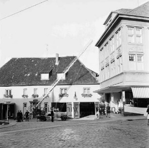 Hude: Feuerwehrgerätehaus "Blaues Haus" mit geöffneten Toren: links Feuerwehrfahrzeug mit ausgefahrener Drehleiter: daran Feuerwehrmänner: rechts Wohn- und Geschäftshaus Gebrüder Petersen und Zuschauer