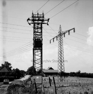 Energieversorgung: Trafostation auf Viehweide: dahinter Strommast der Überlandleitung der Schleswag: links Hauptstraße mit Kopfsteinpflaster und Pkw: im Hintergrund Bauernhäuser