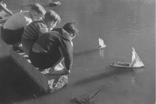 Drei kleine Jungen mit selbstgebastelten Segelbooten, an einem Gewässer spielend