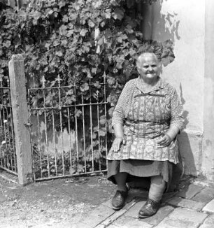 Land Oma : Wasewitz. Eine alte Frau sitzt auf einem Stuhl vor einem Haus, dass mit Wein berangt ist