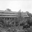 Drahtmühle nach Renovierung: vorn rechts Garten mit Vogelhaus: rechts dahinter zwei Stapel mit Holzbohlen und Brettern: in der Mitte Baum: links Büsche