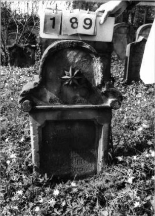 Grabstein 189 (Index: 99792314) Material: Sandstein Zustand: verwittert Maße HxBxT: 77x52x15 cm Beschreibung: Stele, in Voluten auslaufender Rundbogenaufsatz, rechteckiges Schriftfeld eingetieft Symbolik: Voluten, Blüte: Grabstein 189