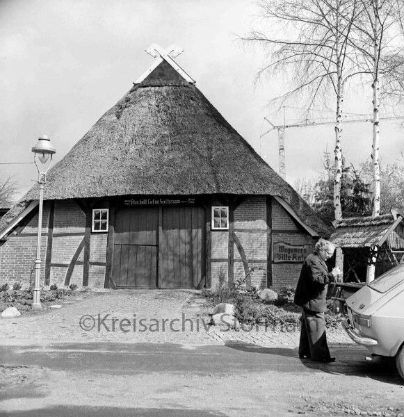 Am Rauchhause: "Wegener's Alte Kate": Giebelfront mit Dielentor und gekreuzten Perdeköpfen als Giebelzier: davor Straßenlaterne: auf Straße Frau Wegner senior bei Pkw: Birken: im Hintergrund Baukran