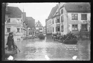 Hochwasser an der Ecke Rechte Tauberstraße/Lindenstraße mit Chorstift-Drogerie, Cafe Quenzer, Eisenhandlung A. Götzelmann u.a. Geschäften in Richtung Brückengasse, darauf Personen auf Schelch sowie Brunnen, darauf Schriftzug "Hochwasser in Wertheim 16. Jan. 1920"