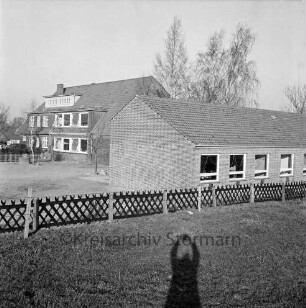 Volksschule: Schultwiete: vorn Rasen mit Jägerzaun: rechts Erweiterungbau: dahinter altes Schulgebäude: hinten Bäume