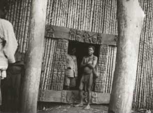 Banssu, Nordwest-Kamerun. Hauseingang mit geschnitztem Türrahmen, davor lendenschurzbekleideter Banso