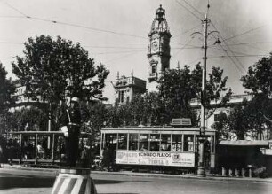 Valencia. Spanien. Straßenszene. Auf der Kreuzung regelt ein Polizist den Verkehr. An einer Straßenbahnhaltestelle steigen Passanten in eine Straßenbahn. Im Hintergrund das Rathaus mit seinem hoch aufragendem Turm