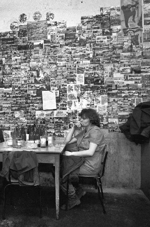 Ansichtskartenwand : Motorenwerkstatt. Arbeiterin sitzt an einem Tisch vor einer Wand mit Ansichtskarten