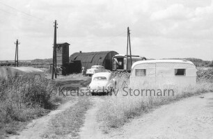 Feldweg am Brenner Moor: Nissenhütte, Campingwagen und Pkws vor Trafohaus und Strommasten: dazwischen Frau und Leine mit Wäsche