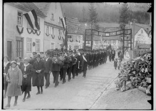 Primizfeier Heinzelmann in Stetten unter Holstein; Prozession; im Mittelpunkt Musikkapelle, dahinter Männer mit Zylinder; im Hintergrund geschmückter Torbogen