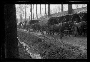 Nicht identifizierbare Ortschaft in Nordfrankreich oder Belgien: Pferdegespanne mit Planwagen auf einer unbefestigten Straße neben einem lang gestreckten Holzschuppen am Rand eines Parks, im Vordergrund ein Bachgraben (Vorderansicht)
