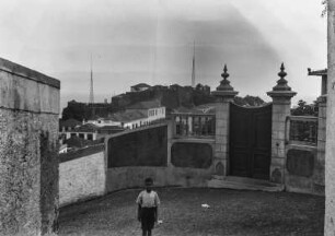 Straßenansicht : Ein Junge steht in einer Straße, hinter ihm eine Mauer mit einem Tor. Im Hintergrund Gebäude und zwei Antennen.