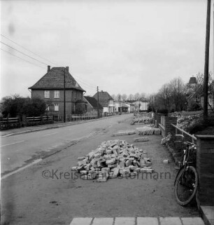 Dorfstraße: Ausbau des Bürgersteigs: Ziegel- und Steinplattenhaufen: vorn rechts Fahrrad: links und hinten Bauernhäuser: im Hintergrund Getreidesilo