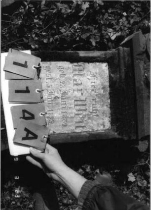 Grabstein 114 (Index: 99792208) Material: Sandstein, weiße Marmorplatte Zustand: verwittert mit Textverlust Maße HxBxT: 110x45x19 cm Beschreibung: Stele, profilierter Sockel, Giebel, rechteckige Schriftplatte Symbolik: Voluten: Grabstein 114A