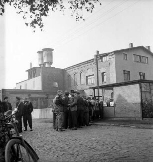 Kampnagel AG: Kampstraße: Eisengießerei: Außenaufnahme: Streik: streikende Arbeiter stehen Schlange vor Pförtnerhaus: hinten Werksgebäude: vorne links Fahrrad [angeschnitten]