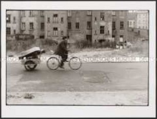 Ein Fahrradfahrer mit Anhänger auf holpriger Straße vor schadhafter Hinterhausfassade (Sonderthema: Anders leben)