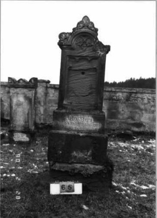 Grabstein 65 (Index: 100114879) Material: Sandstein Zustand: beschädigt am Giebel, sehr stark verwittert am Sockel, sonst leicht verwittert, Textplatte fehlt Maße HxBxT: 238x65x31 cm Beschreibung: Stele auf gestuftem Sockel, eingezogener Rundbogen mit Akroter und Voluten, Blatt im Giebel, eingesetzte Textplatte: Grabstein 65