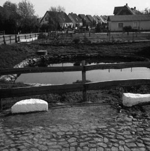 Dorfteich mit Grünanlage und Holzgeländer: davor weiß angemalte Findlinge: hinten Neubausiedlung mit Einfamilienhäusern und Gärten