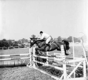 Sportveranstaltung: 2. Pferdeleistungsschau: Veranstalter Reitverein Ahrensburg/Hamburg-Ost: Reiter zu Pferd bei Sprung über Hindernis: Blechtonnen: im Hintergrund Zaun, Häuser