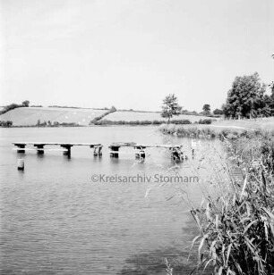 Moorteich mit Schulschwimmsteg: rechts Schilf: hinten rechts Wanderweg und Bäume: im Hintergrund Knicklandschaft