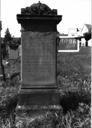 Grabstein 1096 (Index: 100104155) Material: Sandstein Zustand: im oberen und unteren Bereich stark verwittert, sonst noch gut erhalten Maße HxBxT: 138x49x25 cm Beschreibung: Stele auf getrepptem Sockel, auskragender Aufsatz mit Voluten, Mohnkapseln und -blättern Symbolik: Mohnkapseln/-blätter, Voluten: Grabstein 1096