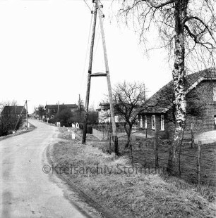 Dorfstraße: vorn rechts Birke und Strommast: dahinter Häuser mit Vorgärten, Zäunen und Hecken: hinten Mitte Kinder