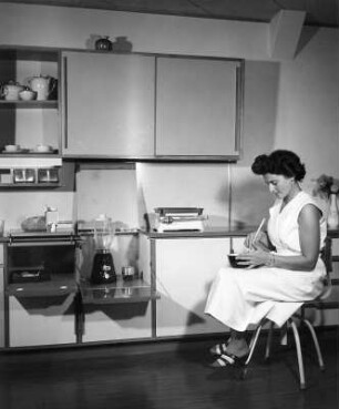 Möbel. Junge Frau, vor einer Anbauküche sitzend und in einer Schüssel rührend