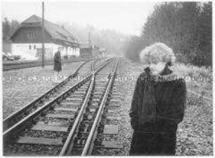 Mädchen steht an Bahngleisen, im Hintergrund ein junger Mann und Gebäude (Altersgruppe bis 14)