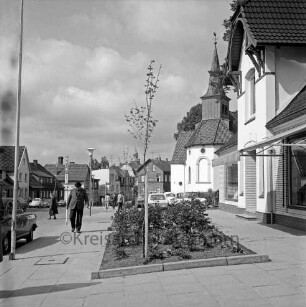 Paul-von-Schoenaich-Straße: Fußgängerweg mit Blumenrabatten: Passanten, Straßenlampen, Verkehrsschilder, parkende Autos: Matthias-Claudius-Kirche: im Hintergrund Bildmitte Rathausturm