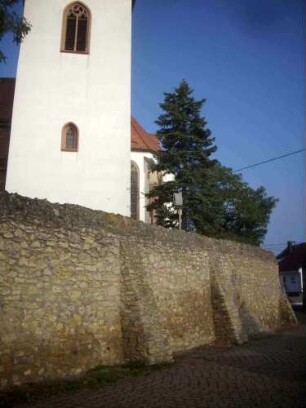 Kirchturm von Süden (Turmkern romanisch - Glockengeschoß spätgotisch erneuert) über Kirchhofmauer (Wehrgang mit Steinbrüstung im 19 Jh abgetragen) mit Strebepfeilern im Verlauf