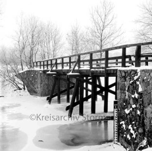 Friedensbrücke: vorn links Trave mit Eisdecke und Schnee: vorn rechts Pegelstandsanzeiger: hinten Bäume: Winter