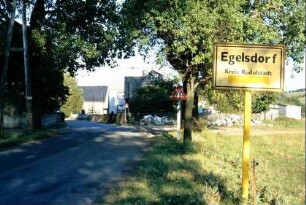 Straße mit Ortsschild, beidseitig Bäume, Häuser
