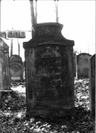 Grabstein 419 (Index: 100115643) Material: Sandstein Zustand: verwittert mit Textverlust Maße HxBxT: 137x171x18 cm Beschreibung: aufgesockelte Stele mit konkav eingezogenem Segmentbogenabschluß, eingetieftes Schriftfeld mit Segmentbogenabschluß Symbolik: Säulen, Blüten: Grabstein 419